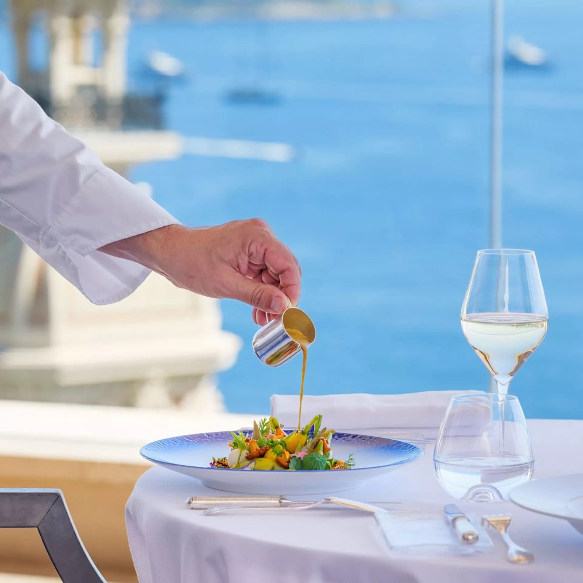 A maître d'hôtel finishes a dish on a terrace, the sea below.